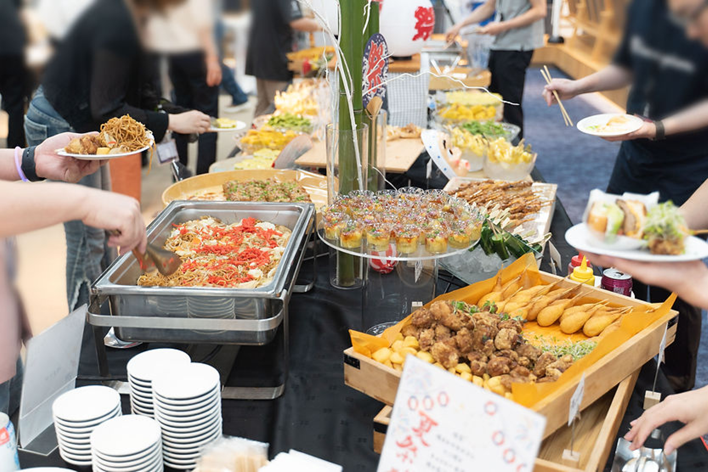 夏祭り風ケータリングで、複数の人が皿に焼きそばや揚げ物、串料理を取り分けている。色鮮やかな料理が並ぶ。