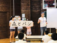 森の国「水際のロッジ」がある愛媛県松野町の小学校にて、地元名産を味わえるパン屋「森とパン」がオープンする話をしている、代表の細羽。児童の皆さんの夏休みの宿題の一つが、「森とパン」の習字。提出をしてもらった中での最優秀賞が、看板ロゴになる、皆さんを巻き込んだプロジェクトです！