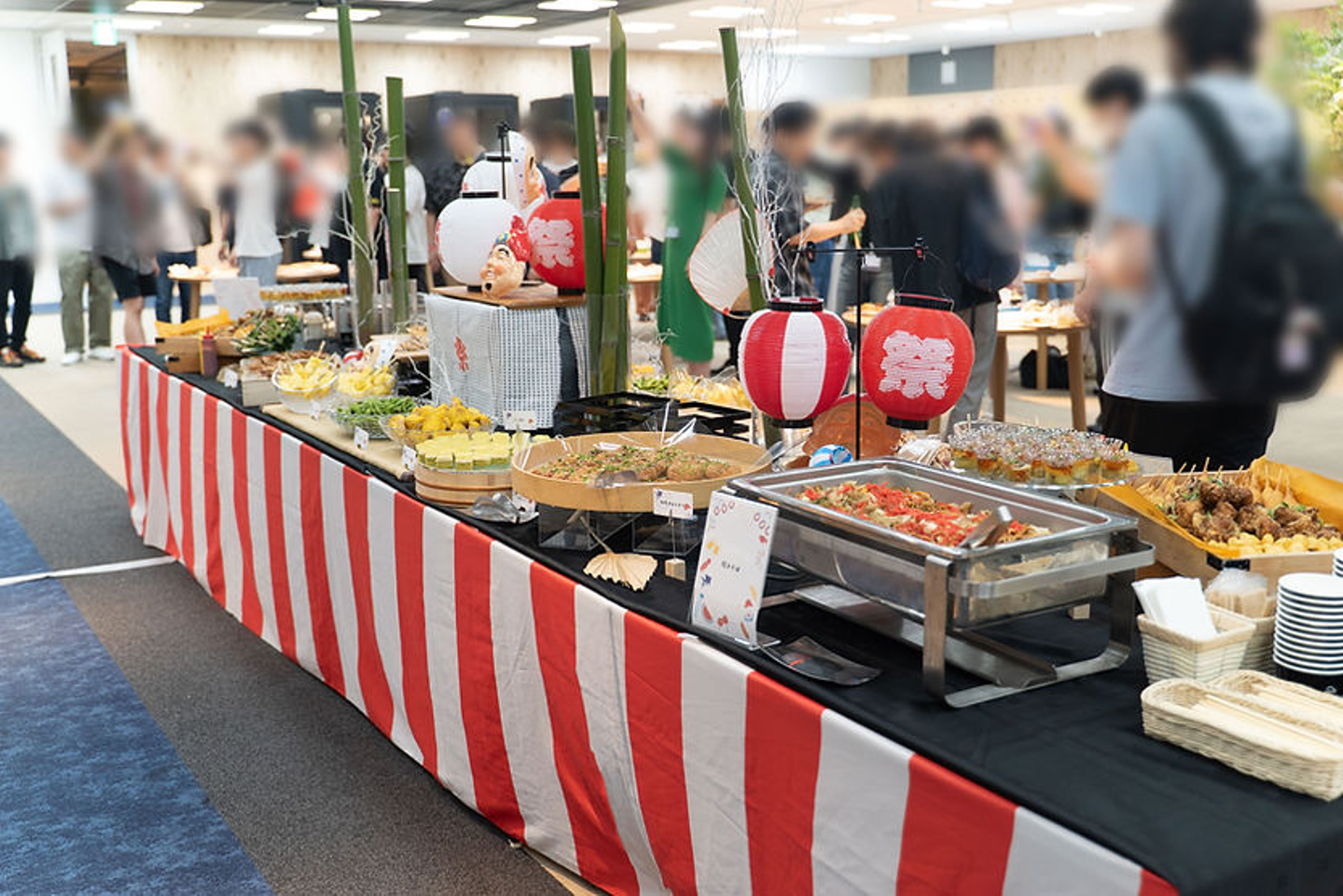 室内の夏祭り風ケータリングで、赤白の提灯や笹飾りの前に料理が並び、多人が集まってにぎやかな雰囲気。