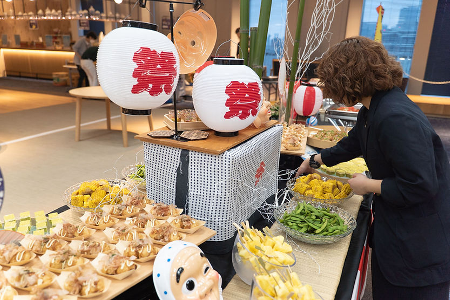 夏祭り風ケータリングで、黒い服のコーディネーターが料理を並べている。串料理やとうもろこし、枝豆、たこ焼きなどが並ぶ。