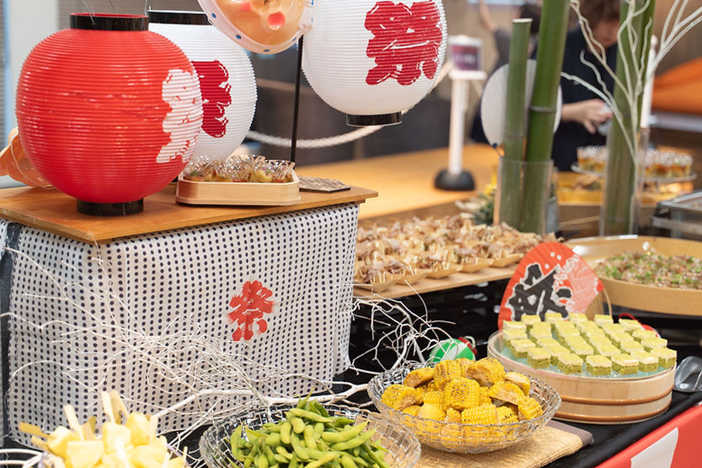 赤白の提灯と竹飾り、手ぬぐいなどがある夏祭り風のケータリング。枝豆、焼きとうもろこし、冷やしパイン、たこ焼き、一口ケーキなどが並ぶ。